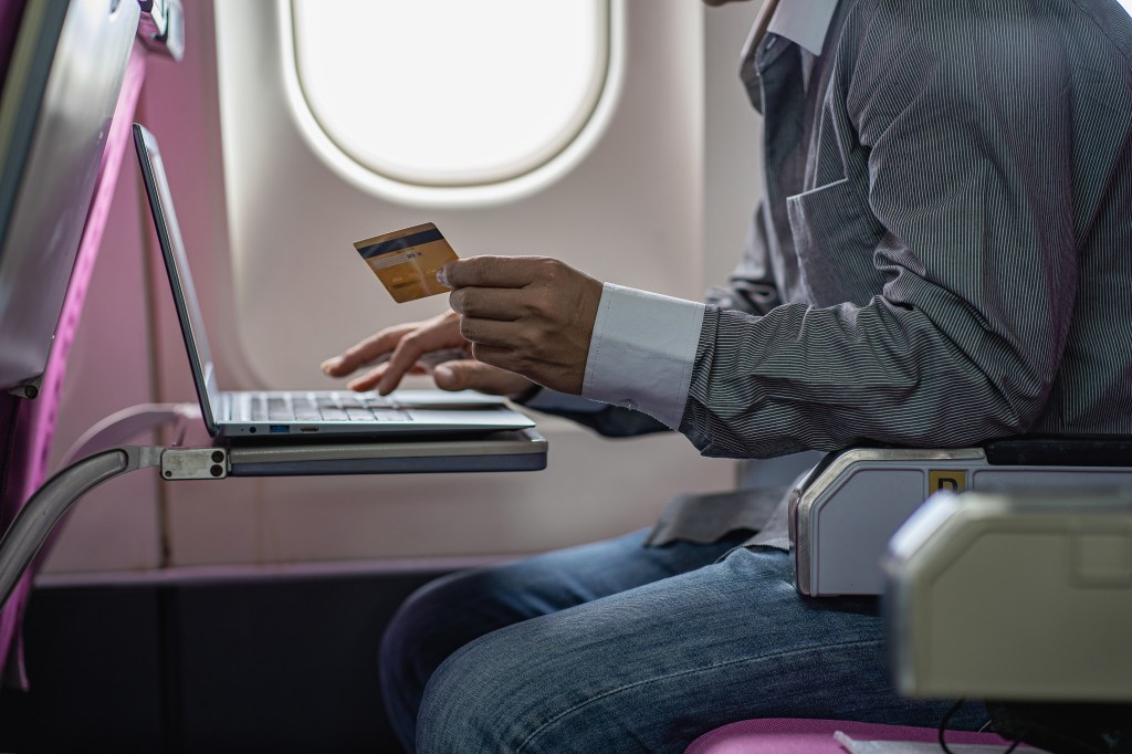 A young Asian man sitting on a plane using a laptop and a credit card for online payments